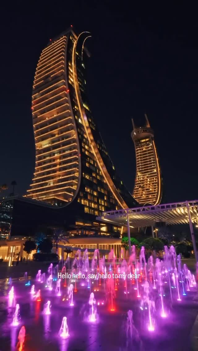 A nighttime shot featuring modern architecture with tall, illuminated buildings.  In the foreground is a fountain with brightly colored lights. The composition is well-lit, with the warm glow of lights against a dark night sky.
