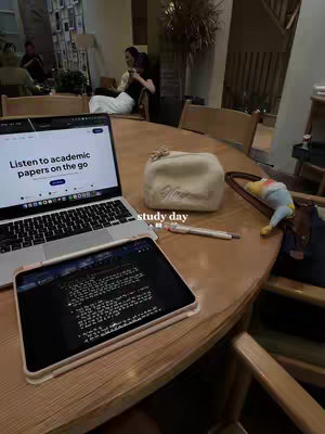 A well-lit photo of workspace in what appears to be a cafe. A laptop on the left, and a tablet in a case are on the tabletop in front, along with a pen and a small makeup bag.