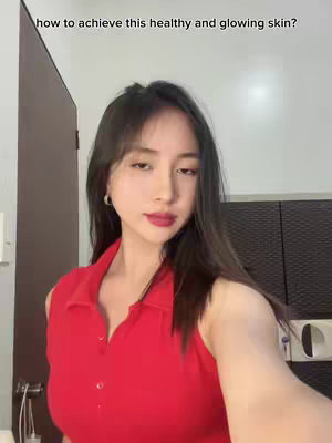 A woman in a red top is shown from the chest up, in what appears to be a selfie video. Her dark hair is long and worn down, and she is looking directly at the camera. Text overlayed near the top of the image asks 'how to achieve this healthy and glowing skin?'. The background is slightly blurred, but includes a wooden door and a partial view of appliances, indicating that the setting appears to be a bathroom or bedroom.