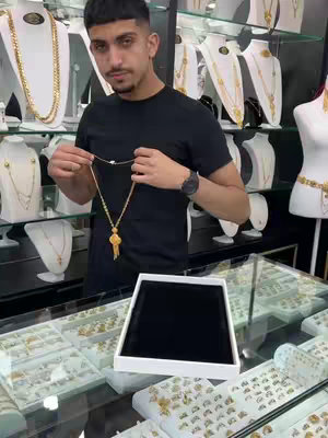 A man in a black t-shirt stands in a jewelry store, holding a gold necklace. Jewelry cases and displays are visible behind him, containing a variety of gold necklaces and jewelry pieces.