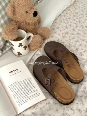 The image showcases a cozy, autumnal scene. Two brown Birkenstock-style clogs rest on a floral-patterned blanket alongside a beige teddy bear holding a small white mug with brown bear illustrations. An open book, with some text visible, and a few other elements add to the inviting vibe. The composition uses soft lighting and warm tones, creating a feeling of comfort and relaxation.