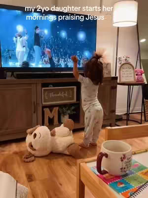 A young girl stands in front of a television displaying a worship service, with a plush dog lying on the floor in front of her and a coffee table to the right with a mug on it.