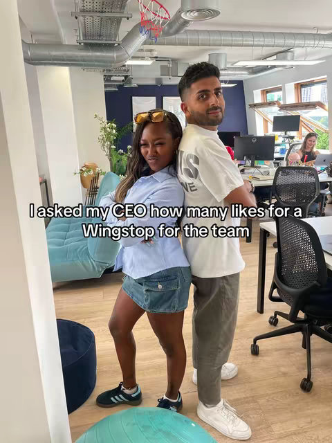 Two people are standing back-to-back in a well-lit office space. The person on the left is a Black woman wearing a light blue collared shirt, a denim mini-skirt, and sneakers. She is wearing sunglasses and smiling. The other person, on the right, is a man with short dark hair, wearing a white t-shirt and grey pants. They both appear to be posing, with the text overlayed at the bottom.