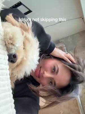 A selfie taken from an angle slightly below the subject, featuring a woman and a dog. The woman has fair skin, reddish blush applied, and brown hair. She's wearing a black top. A fluffy, light-colored dog rests on her chest, its head and nose featured prominently. The background is a minimalistic, possibly indoor setting, with neutral colors. The image is well-lit, with the subjects in clear focus. The woman is smiling, her eyes open and looking at the camera. The hook text is a white color and located on the top right of the frame.