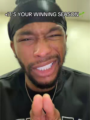 A close-up shot of a man with a black durag, showing a mixed expression of squinting eyes and a slight smile. The man's hands are clasped together in a praying gesture, and he wears a black shirt and a silver necklace. The background appears to be a plain, light-colored wall.