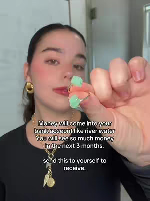 A selfie-style video featuring a woman holding two green rings, likely aventurine, in her hand. She's wearing a black top, gold jewelry, and subtle makeup. Text overlays read, "Money will come into your bank account like river water You will see so much money in the next 3 months. send this to yourself to receive." The background is a muted, neutral-toned interior space.