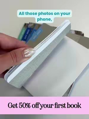 A hand with manicured nails is holding a white phone and opening a white book to reveal pages. The top of the image has the text overlayed with a turquoise banner that says "All those photos on your phone." A pink banner is placed at the bottom of the images with the text"Get 50% off your first book".