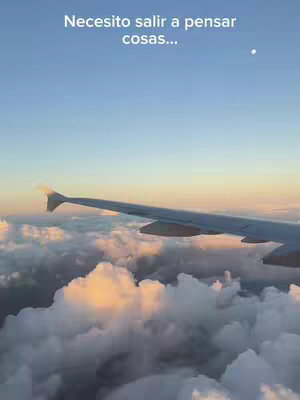 An aerial view from an airplane window, showing a wing against a backdrop of clouds. Above, there's a clear blue sky with the sun setting, casting warm hues on the clouds.