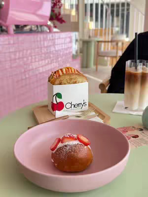 A photo of a cafe scene. A pink frosted counter is in the background with a pink espresso machine. On a light green table in the foreground: a pink bowl holds a pastry topped with strawberries and cream, and another white paper box has 'Cherry's' branding and contains a toasted sandwich.