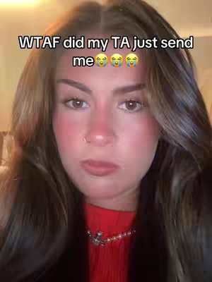 A close-up shot of a young woman with long brown hair, rosy cheeks, and a slight smirk. She's wearing a red top and a silver necklace. The background is dimly lit. Text over the image reads, "WTAF did my TA just send me" followed by crying emojis.