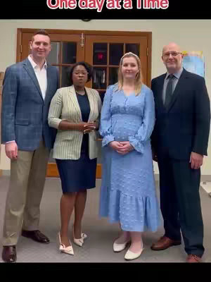 A vertical image showcasing four individuals posing indoors. From left to right, a man is wearing a blue blazer and khaki pants, a woman with short hair is in a navy skirt and a cream colored blazer, a woman in a blue dress, and a man in a navy suit. Behind them is a wooden door.