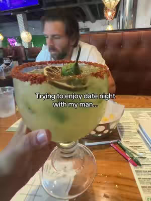 A close-up shot of a person holding a large margarita glass. A man is visible in the background, seemingly looking at something off-camera. The text overlay reads, 'Trying to enjoy date night with my man...'. The setting appears to be a restaurant.