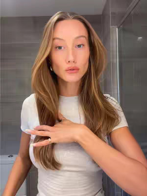 A fair-skinned woman stands in a bathroom, facing the camera with a hand touching her chest. She has long, light brown hair that frames her face,  wearing a white t-shirt. Around her is a neutral-toned bathroom with tiled walls.