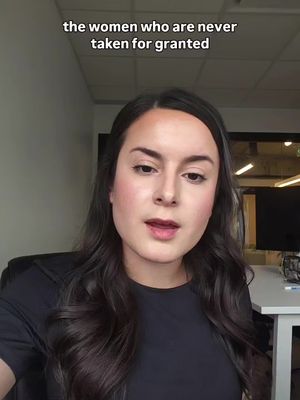 A woman with dark hair and fair skin is centered in a selfie-style shot. She is wearing a black top. The background is a nondescript office setting with neutral walls and some visible office furniture. The image has soft lighting.