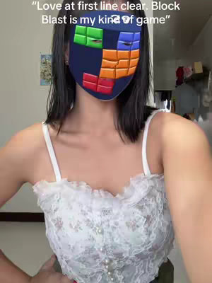 A selfie of a young woman with short dark hair, wearing a white top with floral lace detailing, and a Block Blast game-themed mask. The scene is indoors, with a partially visible wall and a decorative hanging.