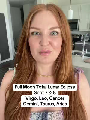 A woman with long red hair and light skin is centered in the frame, looking towards the viewer in front of a kitchen backdrop. Text overlays are present at the bottom displaying astrology information. The woman wears a light-colored top with a decorative neckline and a gold necklace. The kitchen in the background features a white countertop and appliances. The overall atmosphere is well-lit and conversational.