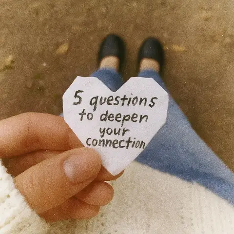 A person holding a heart-shaped paper in front of them, with text written on the paper, and background of legs in jeans. The paper is white. The text reads "5 questions to deepen your connection". The background is blurry.