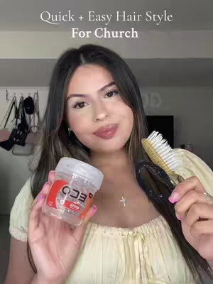 A woman with long, wavy brown hair holds a jar of hair gel, a brush, and a hair clip, smiling at the camera. The background is a simple, neutral-toned room setting. The lighting seems natural and soft.
