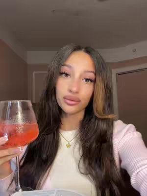 A young woman with long, dark hair and a soft pink top is holding a wine glass filled with a pinkish beverage. She has full lips, prominent eyelashes, and is wearing a delicate gold necklace. The background shows a hotel room with a bed, and the lighting is warm.
