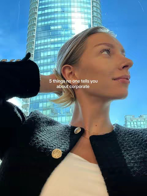 A woman is depicted from the chest up in a slightly low-angle shot. She is fair-skinned with blonde hair pulled back, wearing a black textured jacket over a white top. Gold buttons adorn the jacket. She is looking up at the blue sky with a tall building behind her and text overlay.