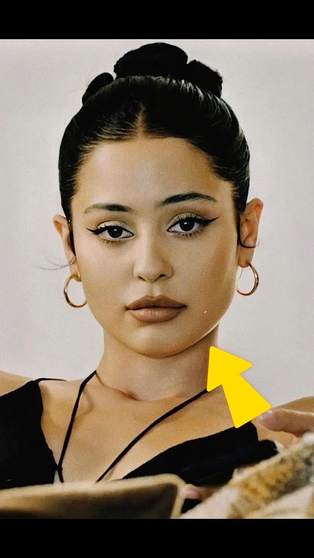 A close-up headshot of a woman with dark hair styled in a bun. She has a fair complexion, bold black eyeliner, and gold hoop earrings. A yellow arrow points towards her shoulder with black attire. The background is a soft, neutral color.