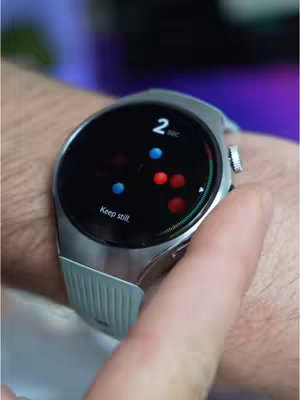 A close-up shot of a person's wrist wearing a silver smartwatch with a light blue band. The watch face is black with a colorful display of small, moving dots of blue and red, and displays the text, 'Keep still.' A finger is touching the watch face.