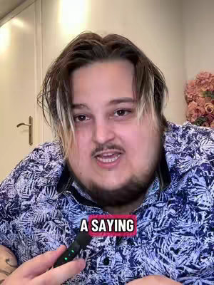 A medium shot features a man in his late 30s or early 40s with light skin, light brown hair and prominent dark facial hair. The man is looking directly at the camera and speaking into a microphone. There is text overlaid on the image saying "A SAYING."
