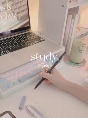 The image showcases a cozy and aesthetically pleasing study setup. The scene is dominated by a silver laptop, with its keyboard prominently displayed, suggesting a focus on work or study. A hand holding a pen is positioned, as if taking notes or journaling. The overall color palette is soft, with a blend of white, beige, and pastel tones, creating a calm and inviting atmosphere.