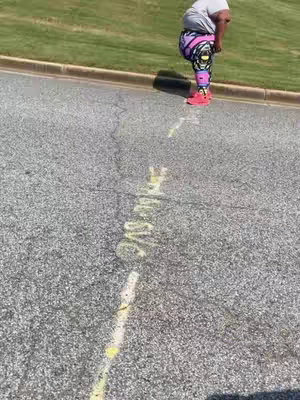 The image shows the lower body of a person wearing colorful leggings and red shoes, appearing to be walking up a slight incline on a textured asphalt road. Yellow painted lines are visible on the road, and green grass borders the asphalt.