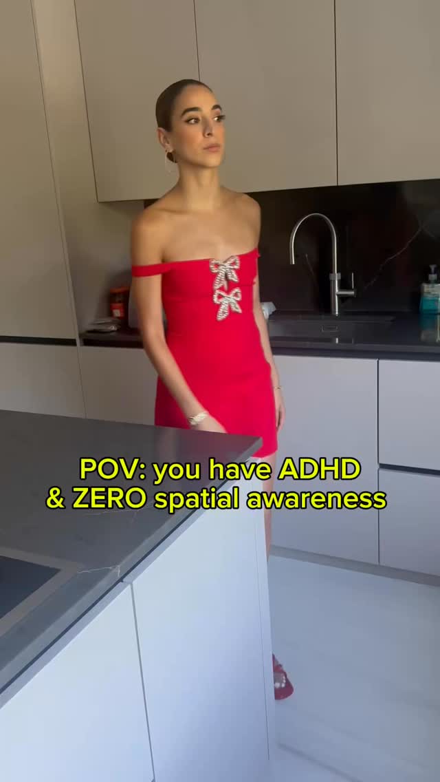 A woman stands in a modern kitchen wearing a red dress. Text overlays state "POV: you have ADHD & ZERO spatial awareness." She has dark hair pulled back and is looking up and to the side.