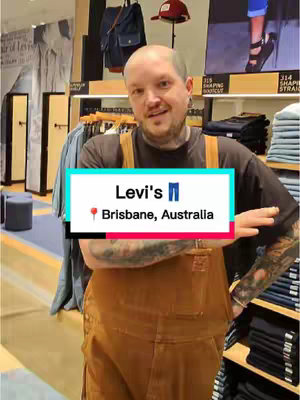 A man stands in a Levi's store, wearing a brown overall and a black t-shirt. A text overlay indicates the location and brand of the store.