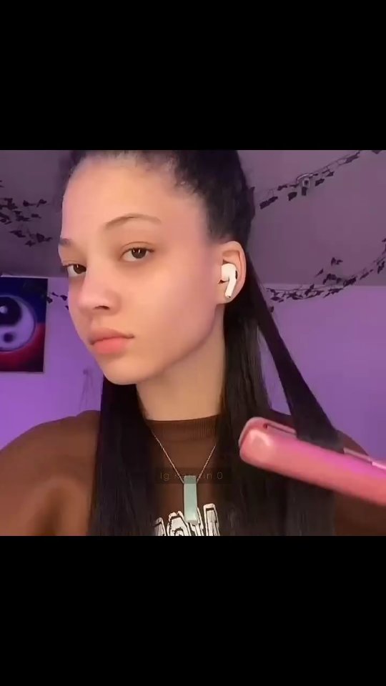 A young woman with dark hair is seen from the chest up, looking towards the viewer with a neutral expression. She is wearing a brown top and has one white earbud in. A piece of her hair is being straightened with a pink hair straightener. The background is dark with purple lighting, and a portion of a wall decor can be seen on the left side.