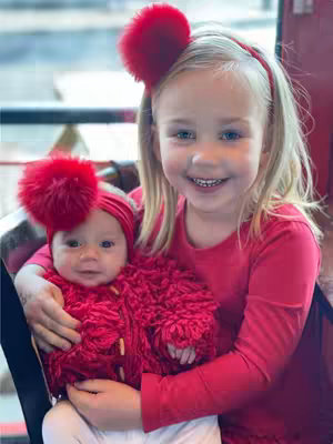 A young girl with blonde hair smiles broadly, holding a baby with a red pom-pom headband and jacket. Both are wearing red outfits, with the background suggesting an indoor setting. The girl is centered, creating a close-up focus on the two subjects.