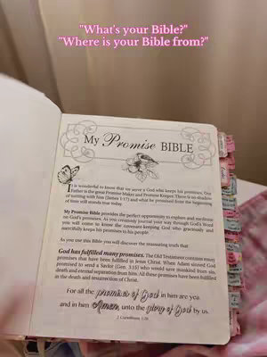An open book is displayed on a surface, likely a bed or soft surface. The book is a Bible, and the cover is visible. The text has a pink tint, and there are various colorful tabs visible on the side of the book's pages. In the background, there's a soft, blurry pink hue suggesting a cozy environment.