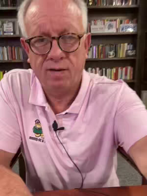 A man with grey hair and glasses is centered in the frame. He is wearing a pink polo shirt with a patch on it. Behind him are bookshelves filled with books. The lighting is natural, and the composition is straightforward, focusing on the speaker.