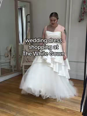 A woman in a white wedding dress is standing in a well-lit room with a large mirror and a chair. The scene suggests a bridal shop or fitting room. The main focus is on the woman in the dress, indicating a wedding dress try-on session.