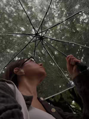 A low-angle shot shows a person holding a clear umbrella in the rain. The person is wearing glasses and a sweater, looking upwards into the umbrella, with a blurred background of green trees. The umbrella's ribs and the rain droplets are visible.