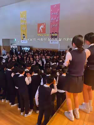 The cover image shows a large group of students in a school hall, with some students on a stage holding microphones. Behind them, there’s a large crowd of students. The setting appears to be an indoor event, and there are banners hanging on the wall.