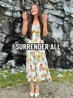 A woman is standing outside, wearing a floral dress, white heels and raising her arms to the sky in worship. A dark rock face is behind her. The words "I SURRENDER ALL" are overlaid on the image.