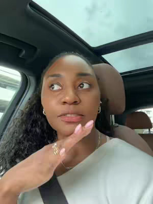 A woman is filming herself inside a car. She has long, dark curly hair. She is wearing a white top and has several rings on, and a necklace. She is gesturing with her right hand and giving a contemplative expression.