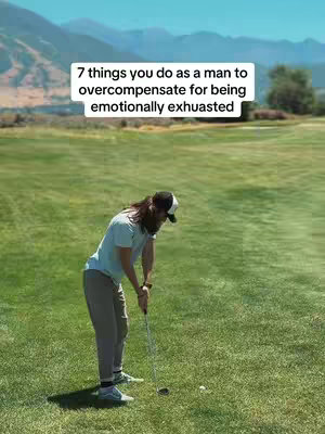 A man in a light blue t-shirt, khaki pants, and a baseball cap is shown in the image, addressing a golf ball on a green field with mountains in the background. The man is holding a golf club, appearing ready to swing. Above him, a rectangular white text overlay details '7 things you do as a man to overcompensate for being emotionally exhauasted.' The scene captures a bright, sunny day, with high contrast.