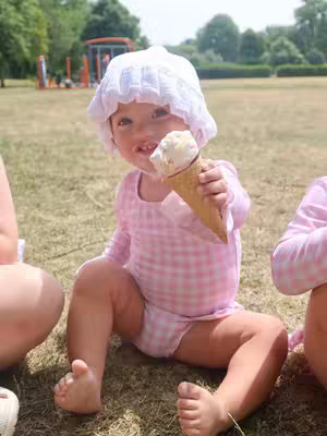 A young child, with fair skin and light-colored hair, is seated on a patch of green grass, holding an ice cream cone. The child is wearing a pink checkered swimsuit and a white bonnet. A playground can be seen in the blurred background, with a sunny atmosphere.