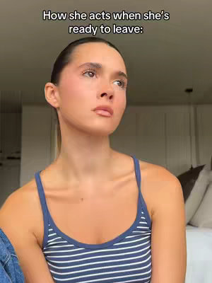 A young woman with fair skin and dark hair is depicted in a medium shot. She's dressed in a blue-and-white striped tank top. The woman's expression is one of subtle emotional strain, with a downward gaze and slightly parted lips. The background includes a blurred interior setting, suggesting a bedroom with a bed and neutral-toned walls.