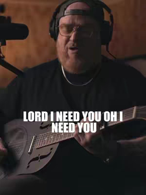 The cover image is a medium shot of a man playing a steel guitar. The man is wearing glasses, a black t-shirt, and a black hat. He also has headphones around his neck. The text overlay "LORD I NEED YOU OH I NEED YOU" is written in a white, bold font. The background has a dark, blurred setting.