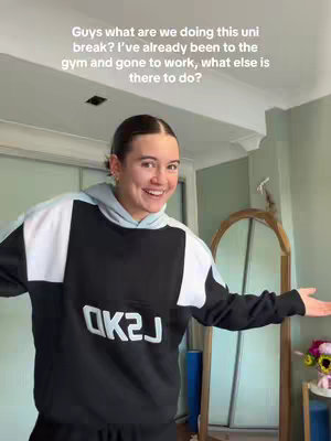A young woman with brunette hair in a high ponytail is standing in front of a mirror. She is smiling and wearing a black, white, and light grey hoodie, and gesturing with her hands. The background includes a room with doors and a mirror.
