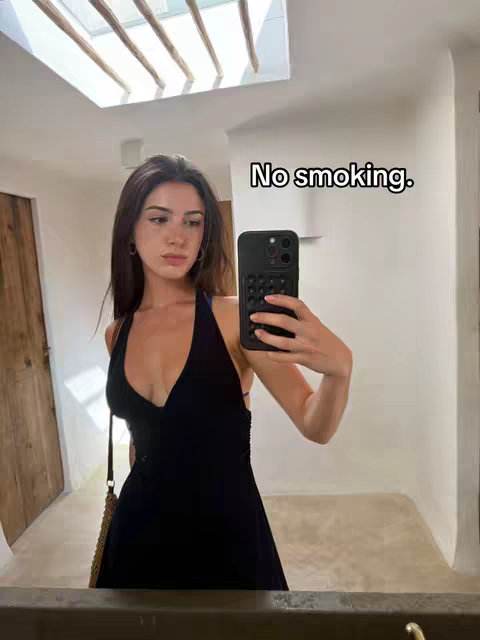 A woman takes a selfie in a mirror. She is wearing a black dress and has long brown hair. She is holding her phone in front of her face. The text overlay reads "No smoking."