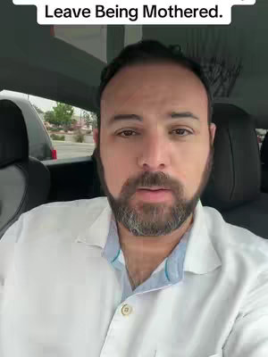 A man in a white shirt is centered in the frame, seated inside a car. The shot is framed from the chest up, with the man's face as the primary focus. His expression is neutral. The car interior includes visible door panels and the front seat. A portion of the outside environment, including parts of other vehicles.