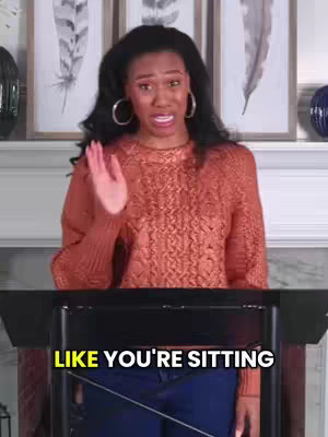 A woman with long, dark hair and large hoop earrings speaks passionately, gesturing with her right hand. She wears a rust-colored sweater with a cable knit pattern. Behind her, a wall is adorned with framed feather artworks. The text overlayed at the bottom gives context to her speech.