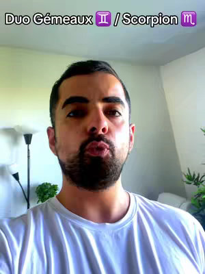 A man with a short beard is in the center of the frame, pouting his lips slightly. Above his head, text reads 'Duo Gémeaux ♊ / Scorpion ♏'. The background appears to be a bright room with white walls and a light-colored ceiling. There are soft shadows and the overall lighting appears to be natural.