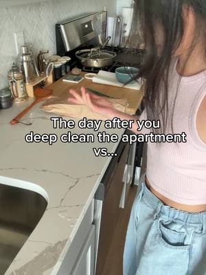 A woman in a pink tank top and light blue jeans is standing in a kitchen. Text on the image reads, "The day after you deep clean the apartment vs..." The kitchen features a stainless steel stove, a white countertop, and various kitchen items.